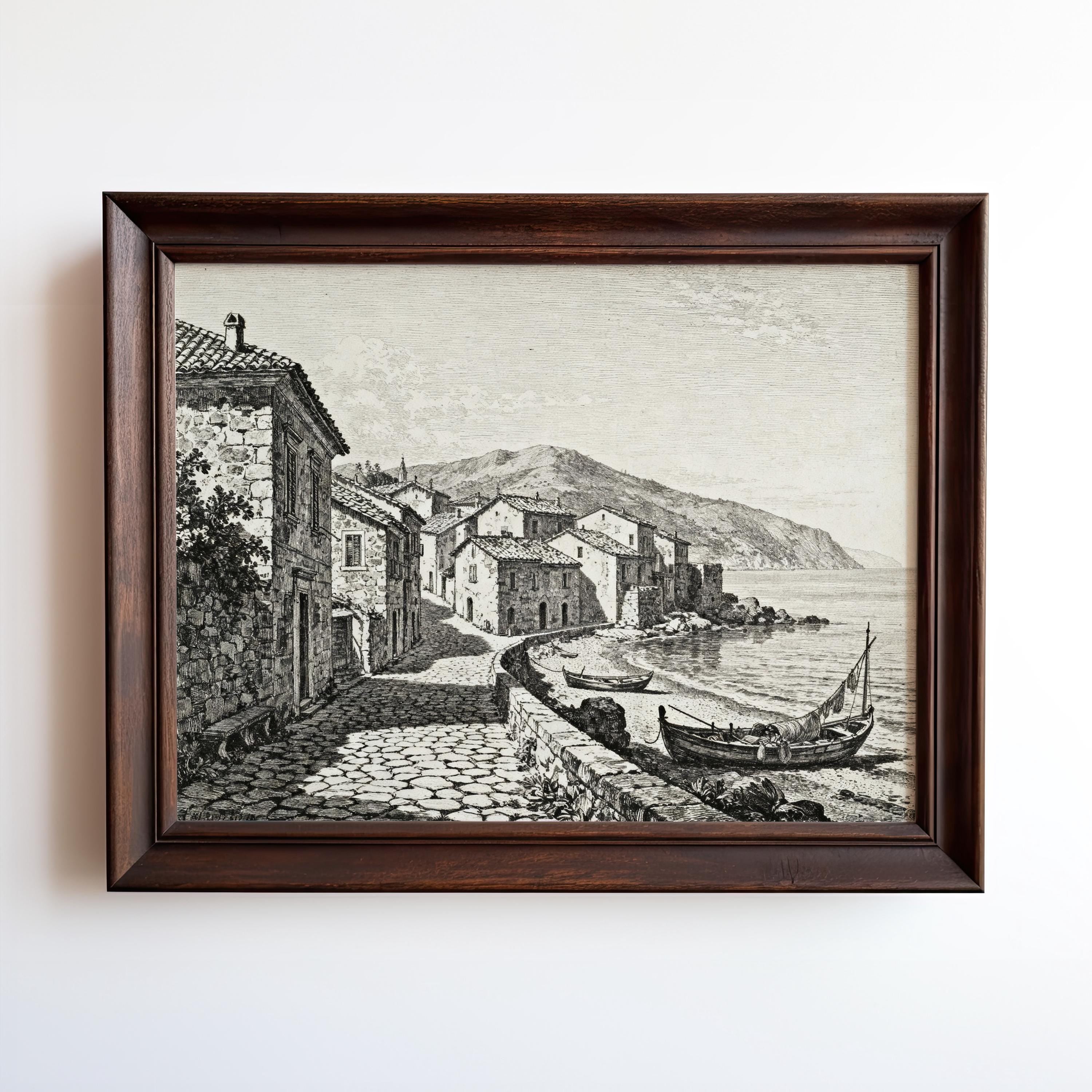 The Coastal Village Art Print is a vintage seaside wall art featuring a black and white etching of stone buildings along a cobblestone path, boats by the shore, and mountains in the background under a lightly clouded sky.