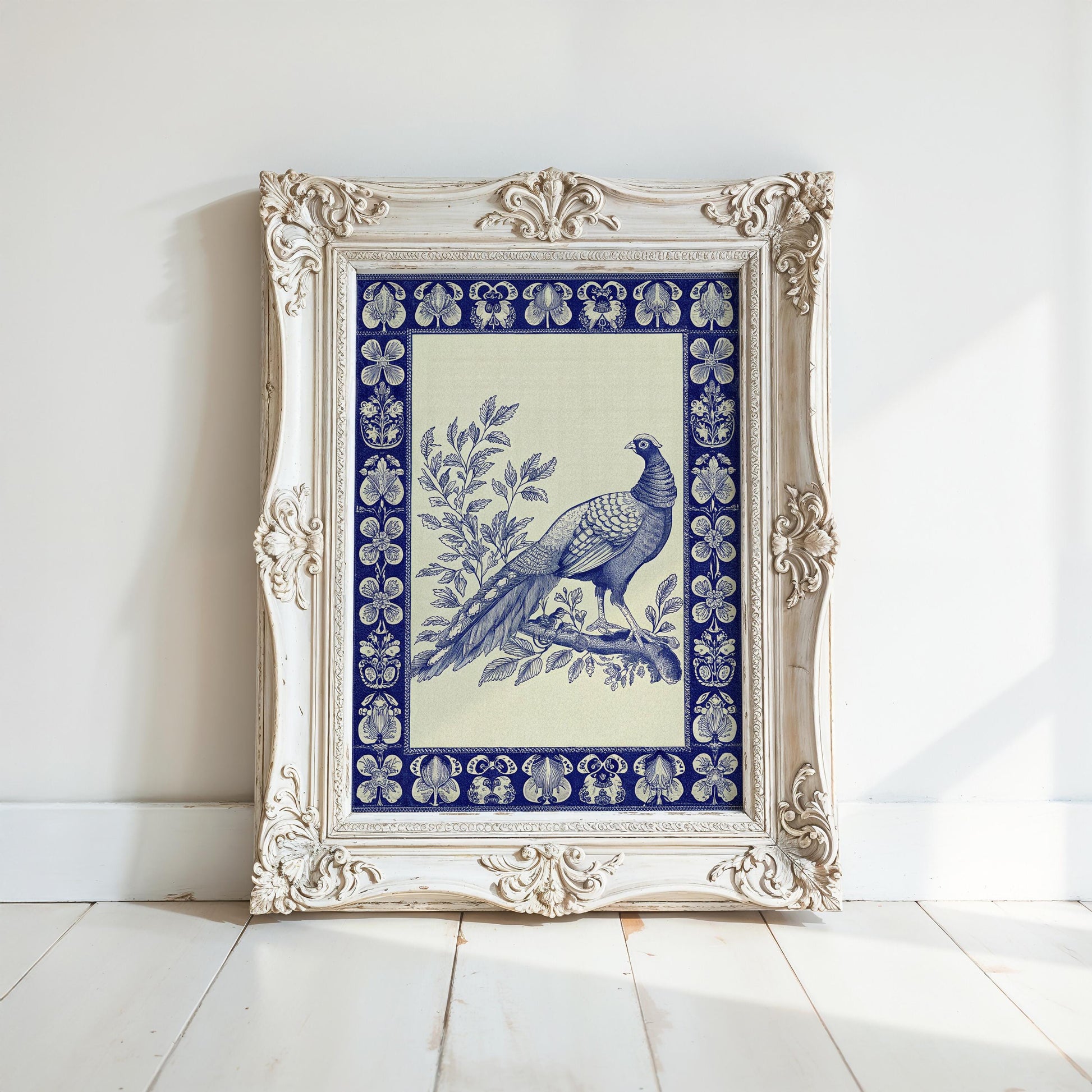 A Vintage Pheasant Wall Art print—an indigo bird with a blue border in ornate wood and metal frames—leans against a white wall on a light wooden floor.
