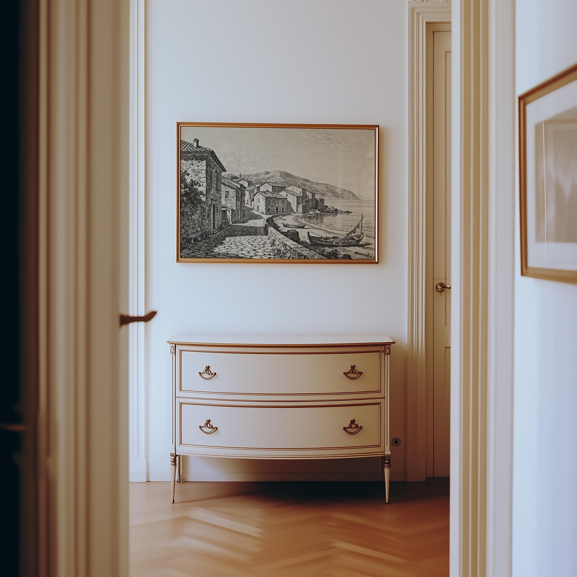 A cream-colored dresser with gold accents sits against a white wall, beneath the Coastal Village Art Print—vintage seaside wall art in black and white—creating a timeless focal point in the bright hallway with wooden parquet flooring.