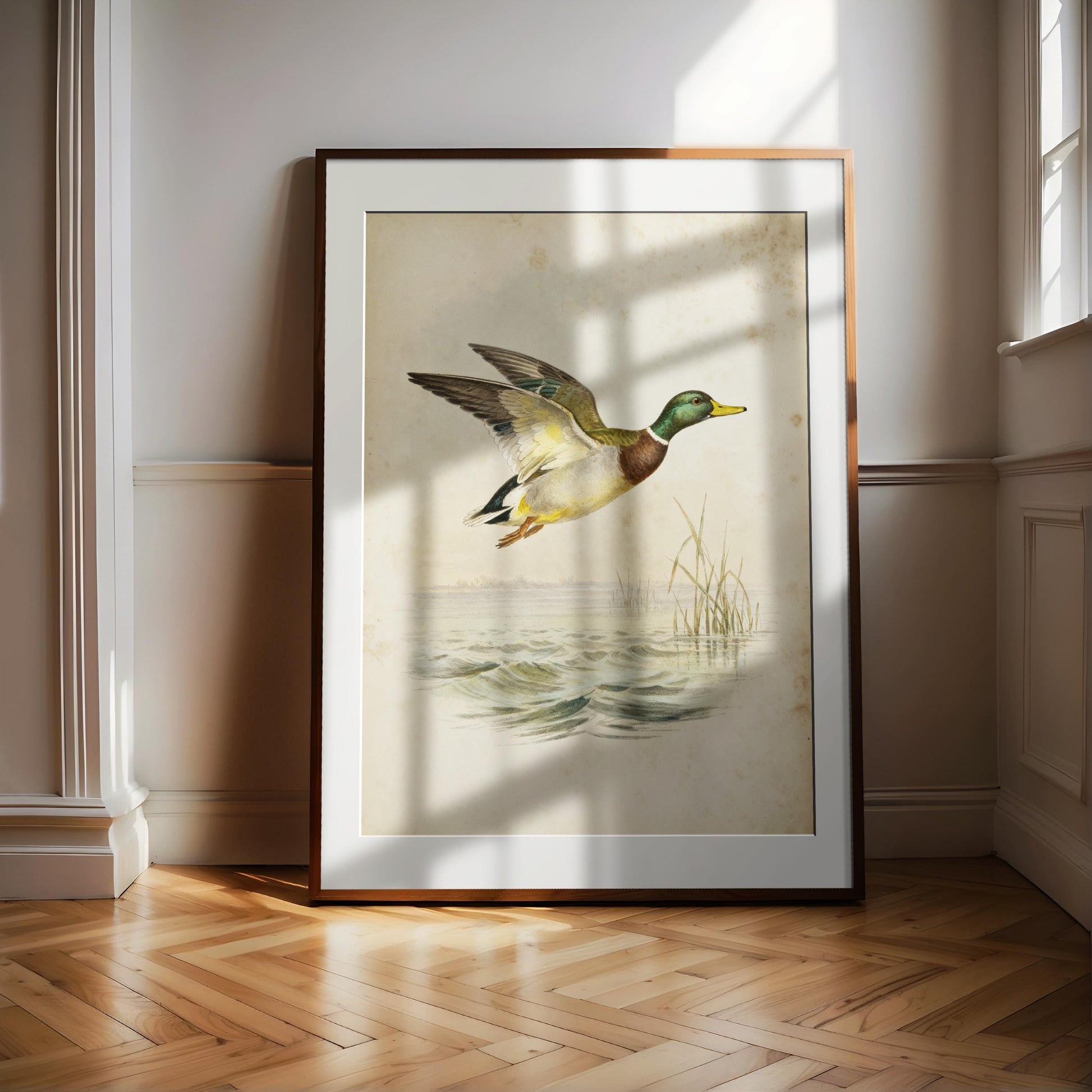 A framed painting of a duck in mid-flight, with a serene body of water and reeds in the background, is displayed on a wooden floor in a room with white walls.
