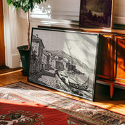 The Coastal Village Art Print – Vintage Seaside Wall Art, a black-and-white etching in a frame, leans against a wooden cabinet near a potted plant on a hardwood floor with a red patterned rug, sunlight streaming in from the left.