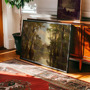 The Vintage Swamp Landscape Print – Cypress Tree Art, a framed nature print from the West & Wild Art Archive, leans against a wooden cabinet on hardwood flooring near a patterned rug and potted plant in a sunlit room.