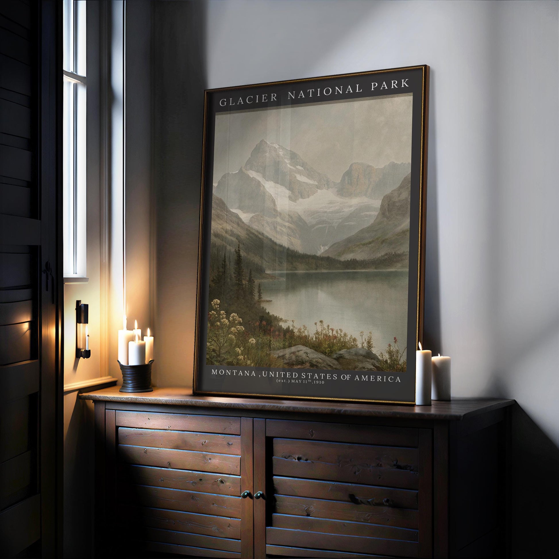 A Glacier National Park Print – Vintage Mountain Landscape Poster with wood or metal frame rests on a sideboard near a window, surrounded by lit candles. The vintage art print shows mountains, a lake, and wildflowers in archival quality.