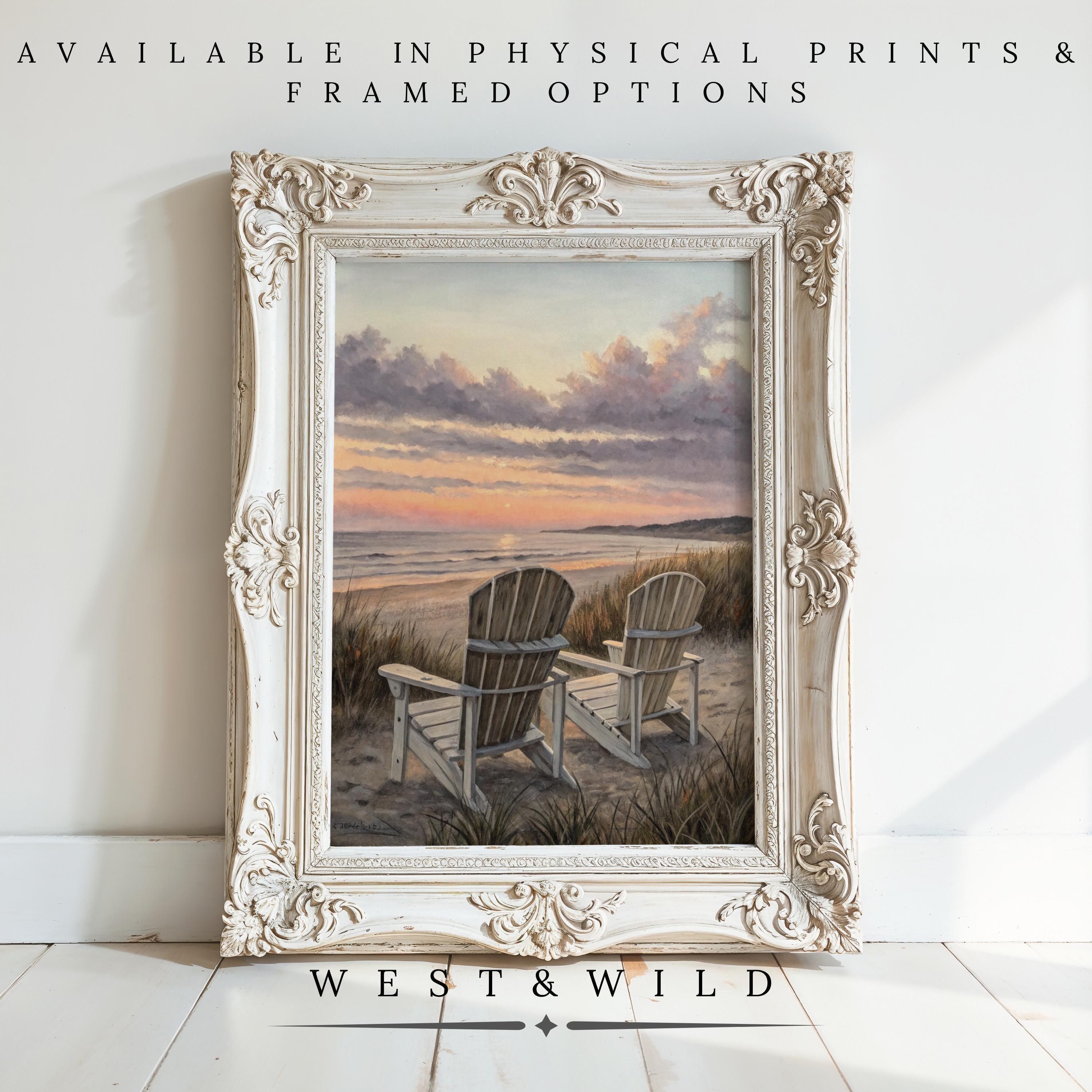 An ornate white frame holds a beach sunset painting of two empty chairs facing the ocean, leaning against a white wall. Beach Sunset Painting – Coastal Wall Art with framing options available from WEST&WILD.