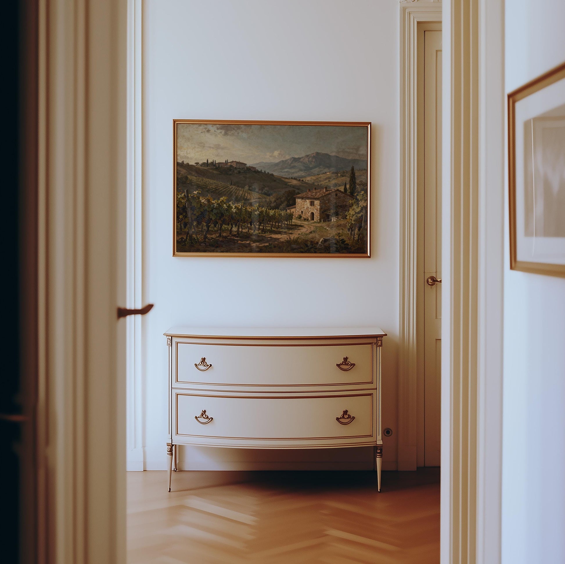 A hallway with light walls and wood floors features a classic white dresser with gold handles beneath the Tuscan Vineyard Art Print – Framed Farmhouse Decor, adding Mediterranean style to the space.