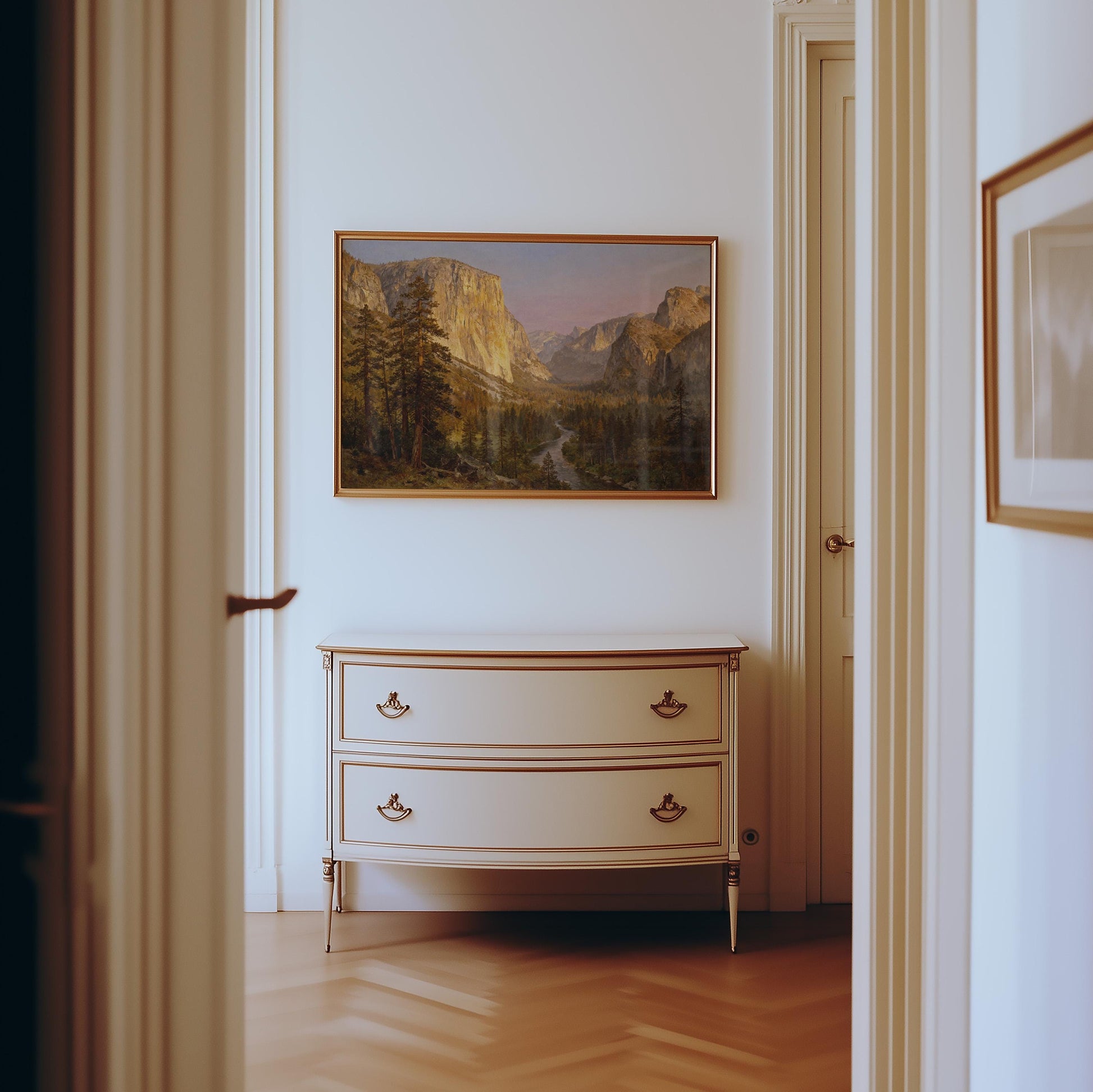 a hallway with a painting of a mountain landscape hanging on the wall above a white dresser with two drawers.