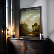 The Everglades Vintage Poster – Florida Wall Art | Coastal Landscape Print is displayed framed on a wooden cabinet with lit candles, creating a cozy atmosphere next to a window.
