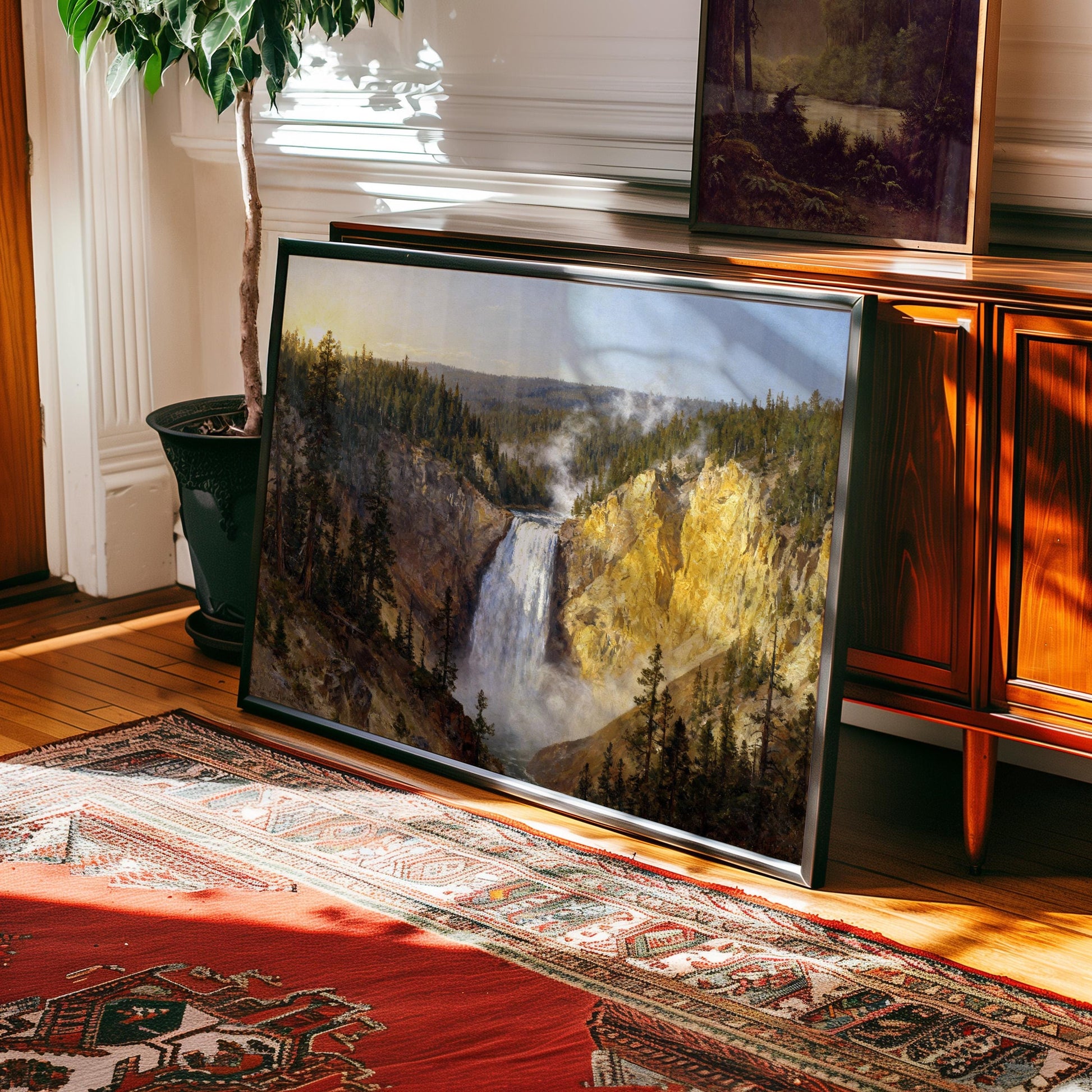a large framed painting of a waterfall, placed on a rug in a room with a wooden cabinet and a potted plant.