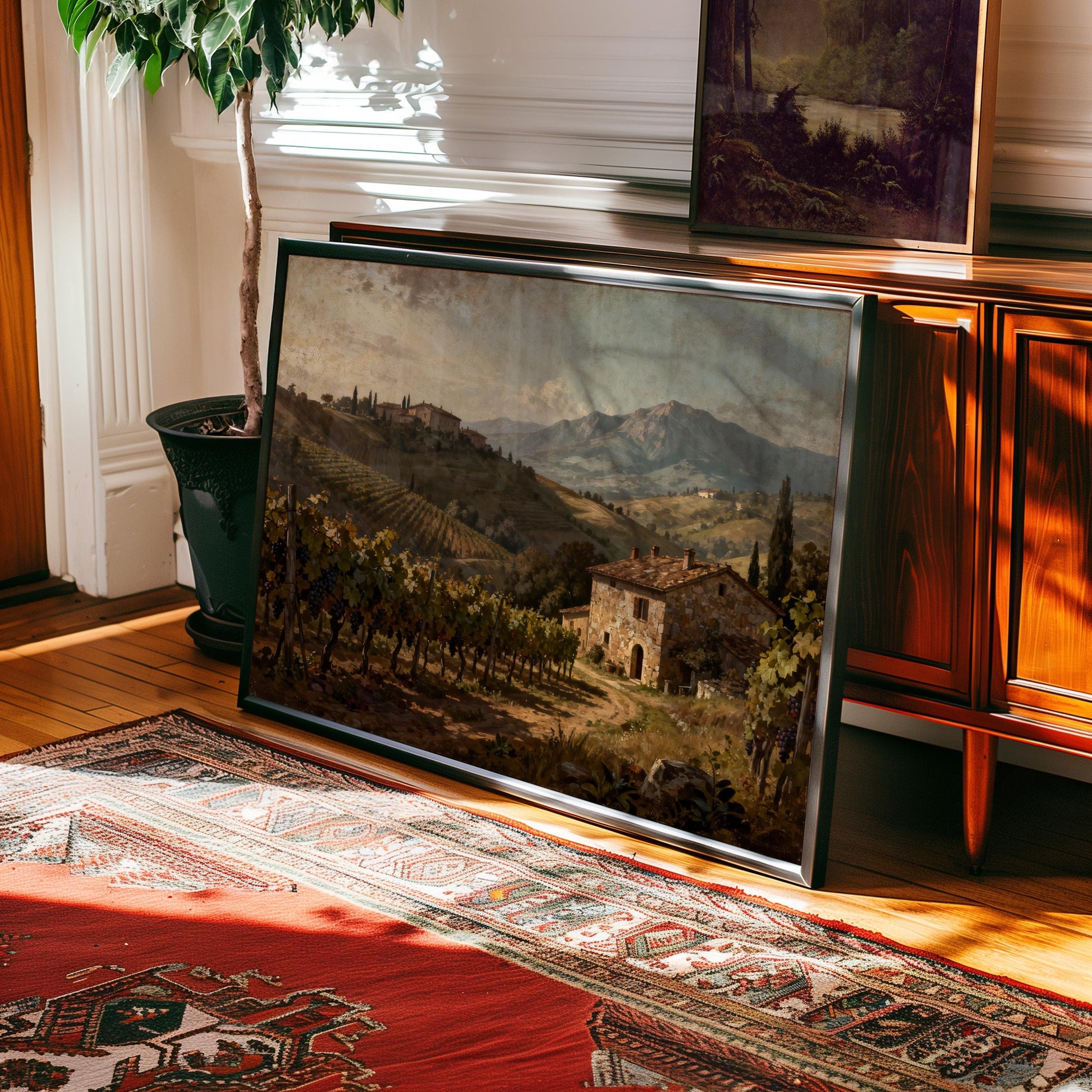 The Tuscan Vineyard Art Print – Framed Farmhouse Decor features a rustic stone house and village, displayed on a wood console beside a potted plant in a sunlit room—ideal for Mediterranean-inspired home decor.
