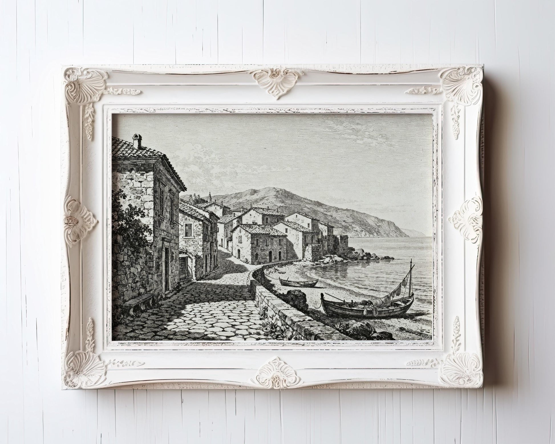 The Coastal Village Art Print is a vintage seaside wall art piece showing coastal houses, boats by the shore, and mountains in black and white etching—featured in an ornate white frame with decorative corners.