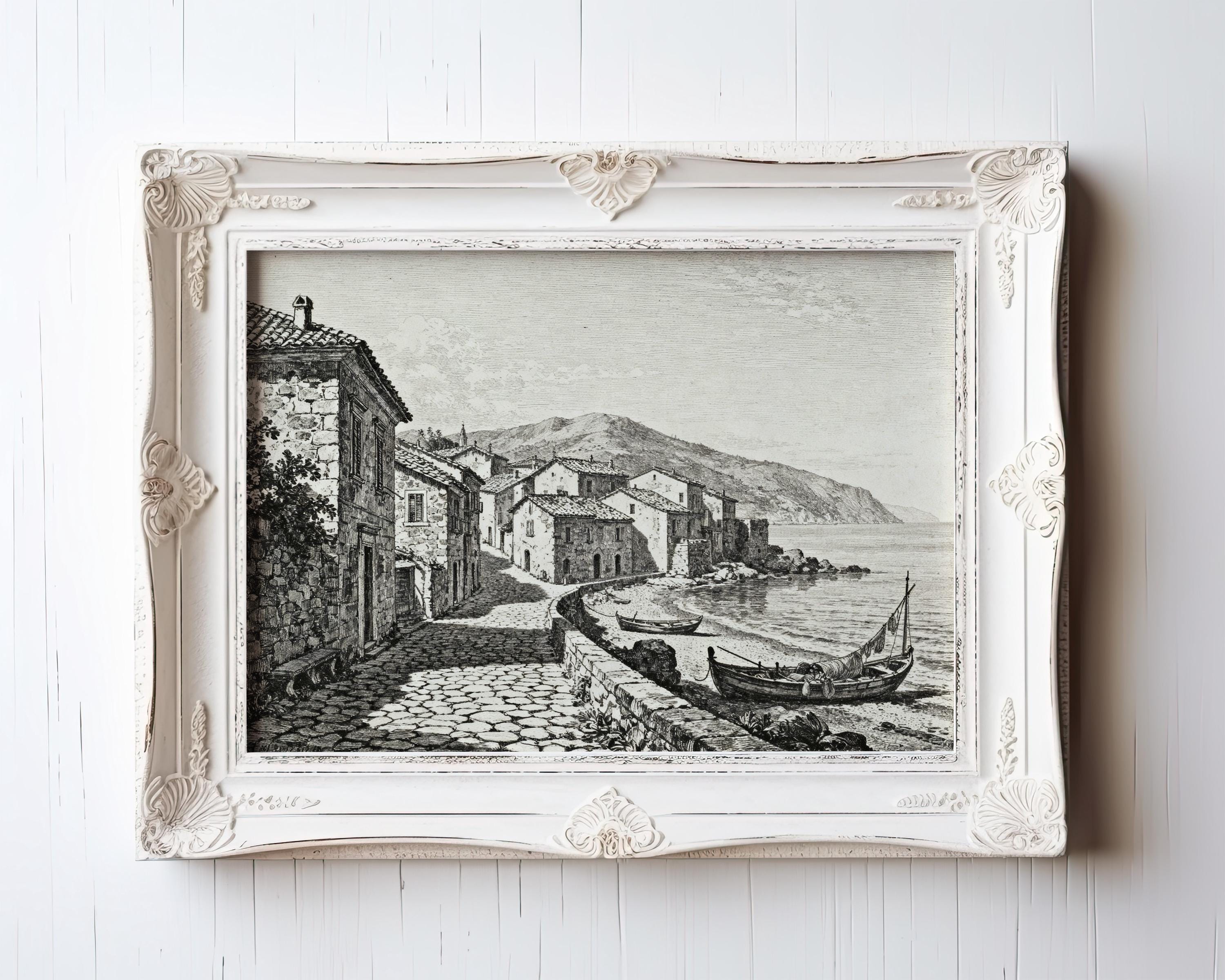 The Coastal Village Art Print is a vintage seaside wall art piece showing coastal houses, boats by the shore, and mountains in black and white etching—featured in an ornate white frame with decorative corners.