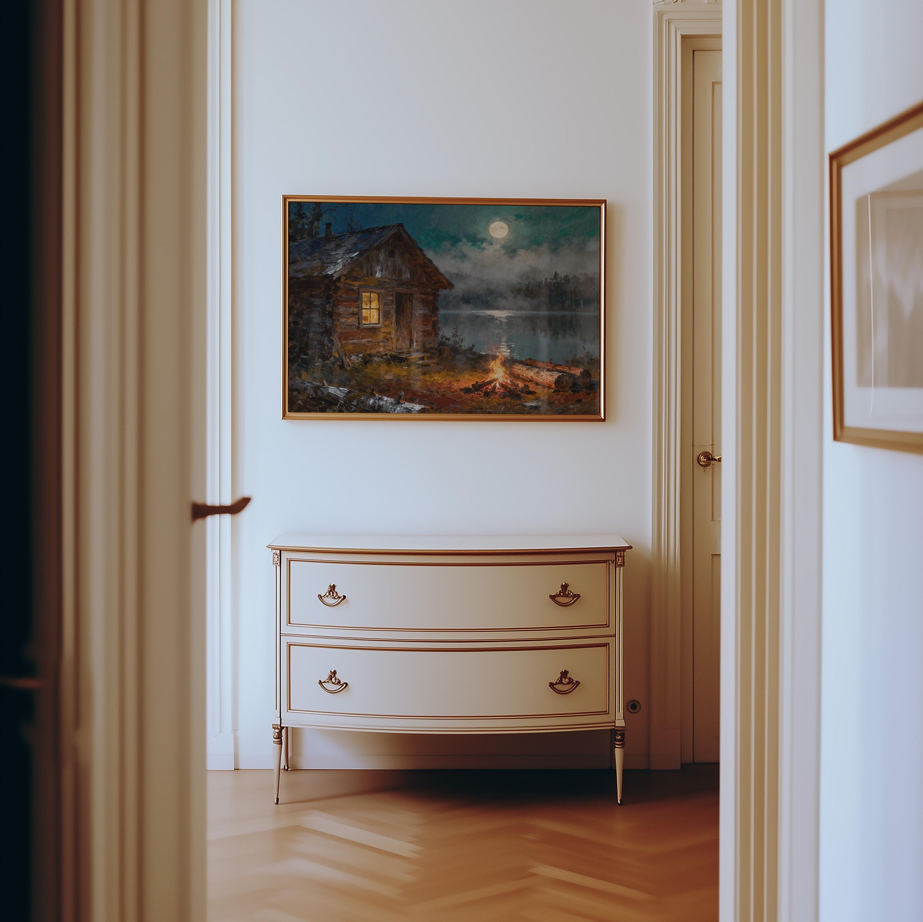 A hallway with light walls and wood floor features a cream dresser with gold handles. Above it hangs the Vintage Log Cabin Print—Rustic Cabin Art with Campfire Glow & Moonlit Lake from Vintage Art Prints & Framed Art.