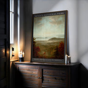 A framed Acadia National Park Poster – Vintage Botanical Coastal Print with Sand Beach Illustration sits on a wooden cabinet next to three lit candles, sunlight streaming in. The art print depicts a scenic landscape of trees and distant hills.