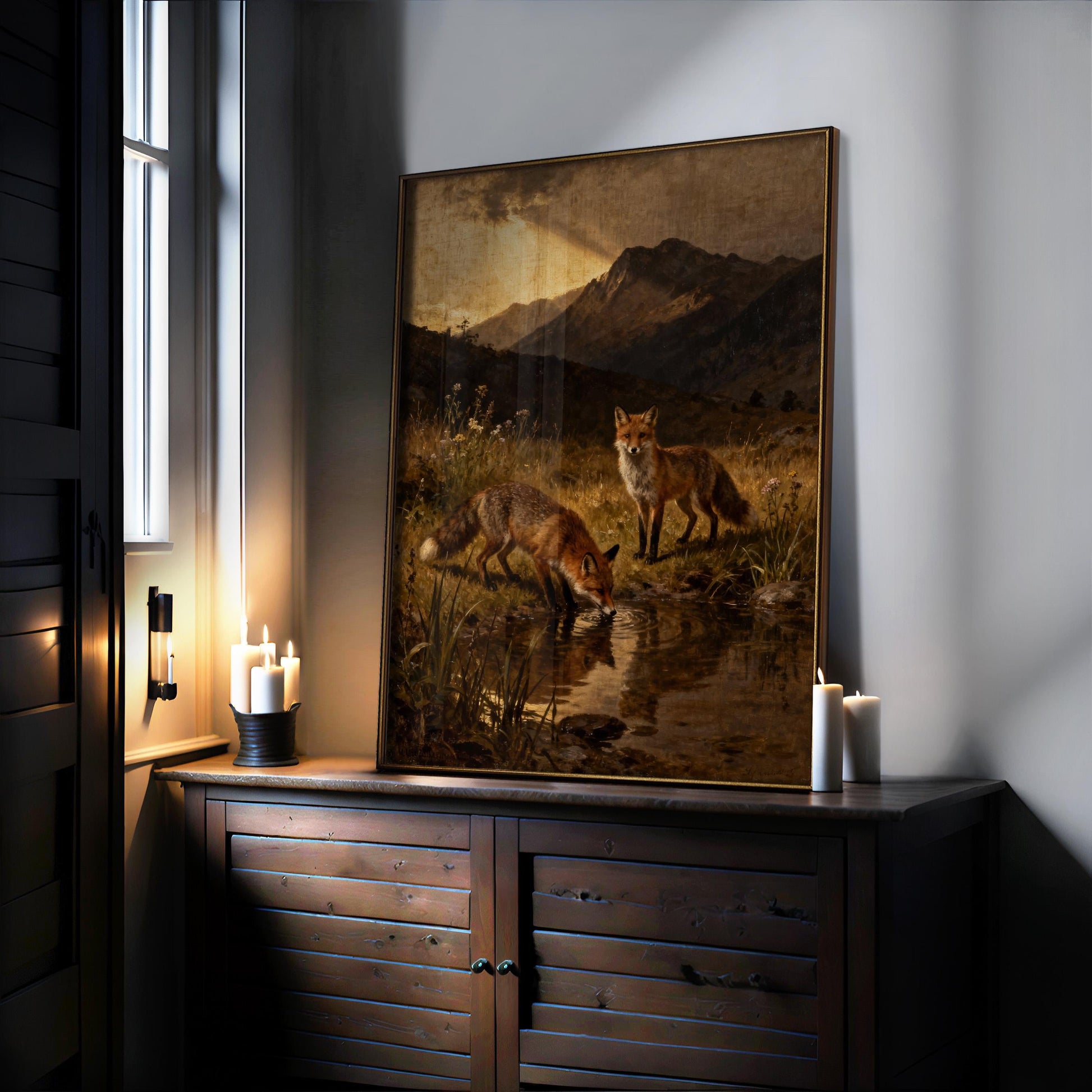 a painting of two foxes in a grassy field, with one fox drinking from a pond and the other standing nearby. The painting is displayed on a wooden cabinet or shelf, with candles and a window in the background.