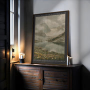 The Rocky Mountain Print – Framed Botanical Wall Art sits on a wooden cabinet beside lit candles, showcasing mountains, a lake, and rocky terrain. Soft light filters in nearby, highlighting this inviting piece of mountain-inspired décor.