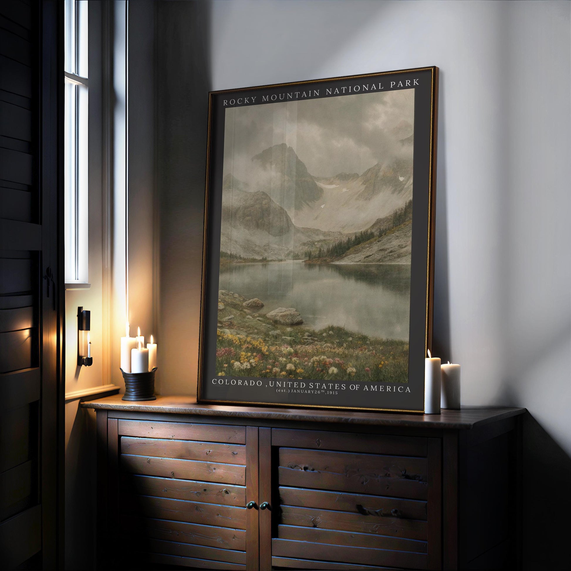 The Rocky Mountain Print – Framed Botanical Wall Art sits on a wooden cabinet beside lit candles, showcasing mountains, a lake, and rocky terrain. Soft light filters in nearby, highlighting this inviting piece of mountain-inspired décor.