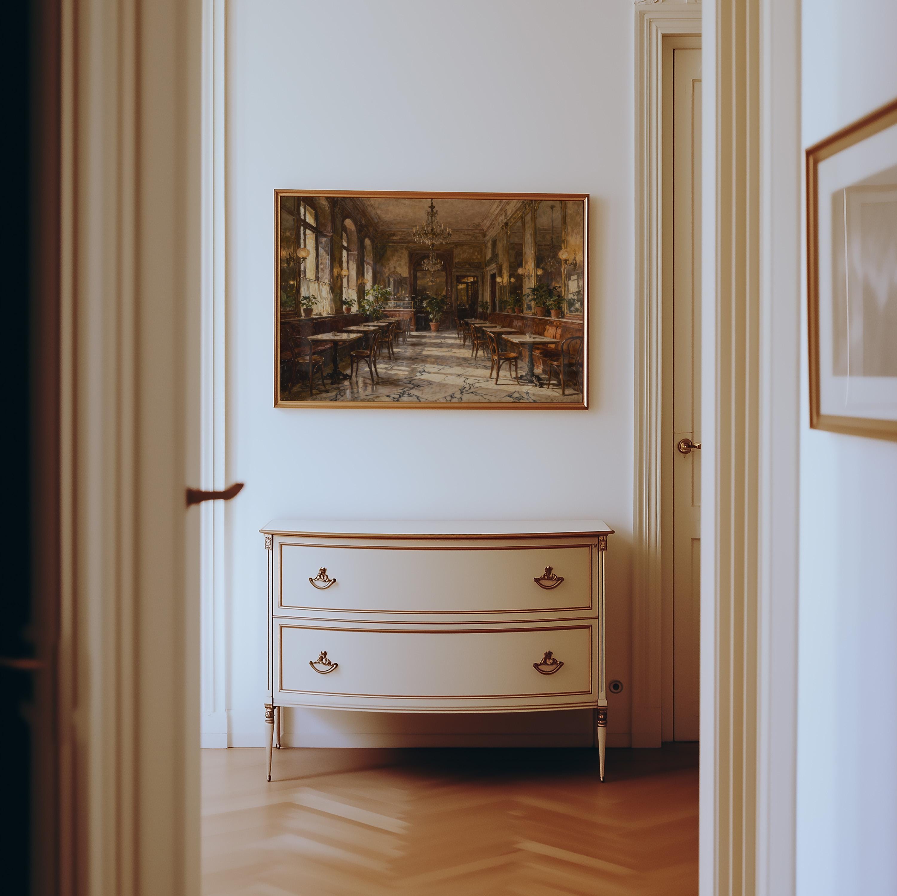 A cream-colored dresser with gold accents stands against a white wall, beneath a framed Vintage European Cafe Print featuring classic marble floors, warm light, and chandeliers from West & Wild Art Archive.