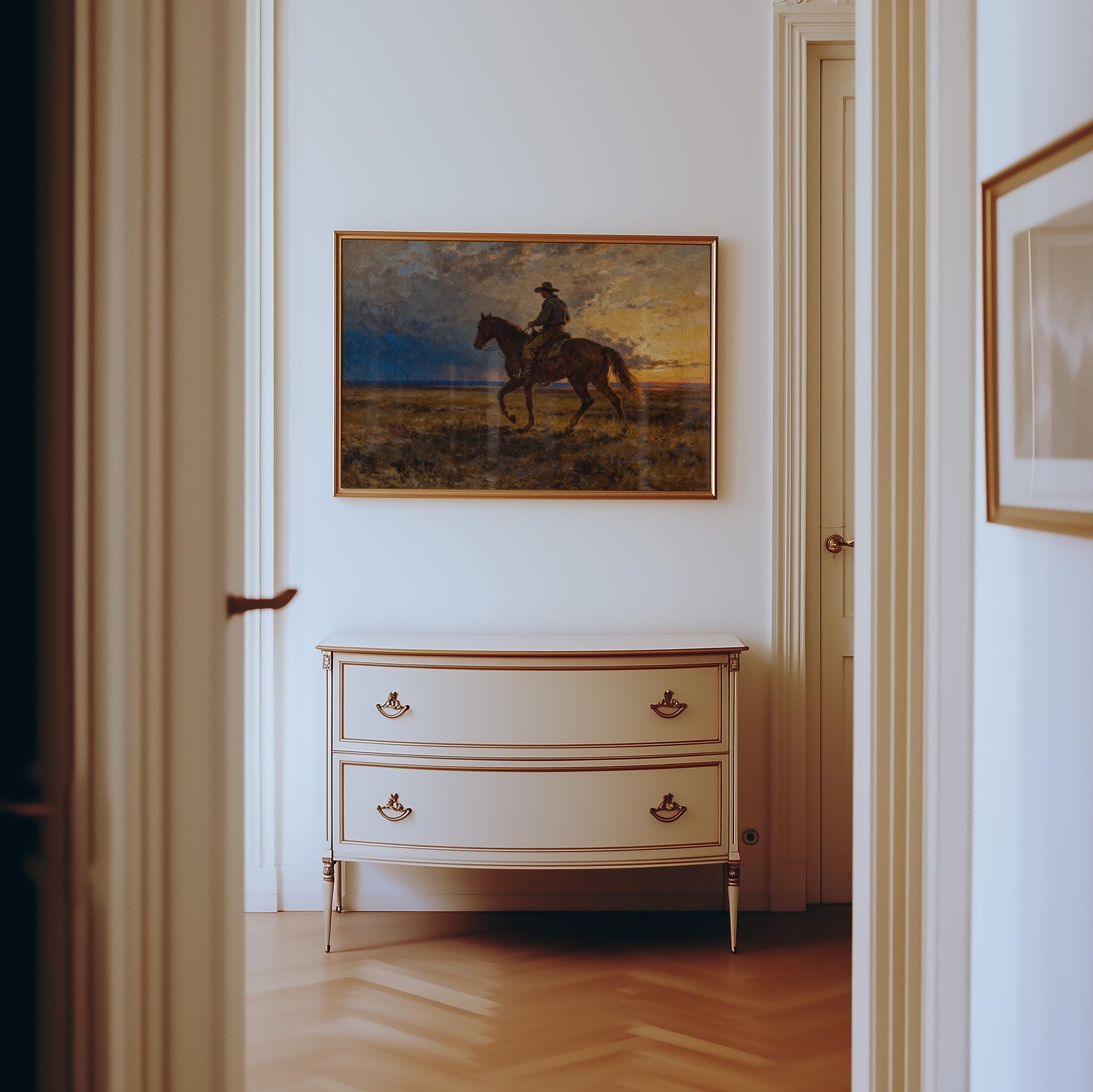 a hallway with a painting of a cowboy on a horse hanging on the wall above a white dresser with two drawers.