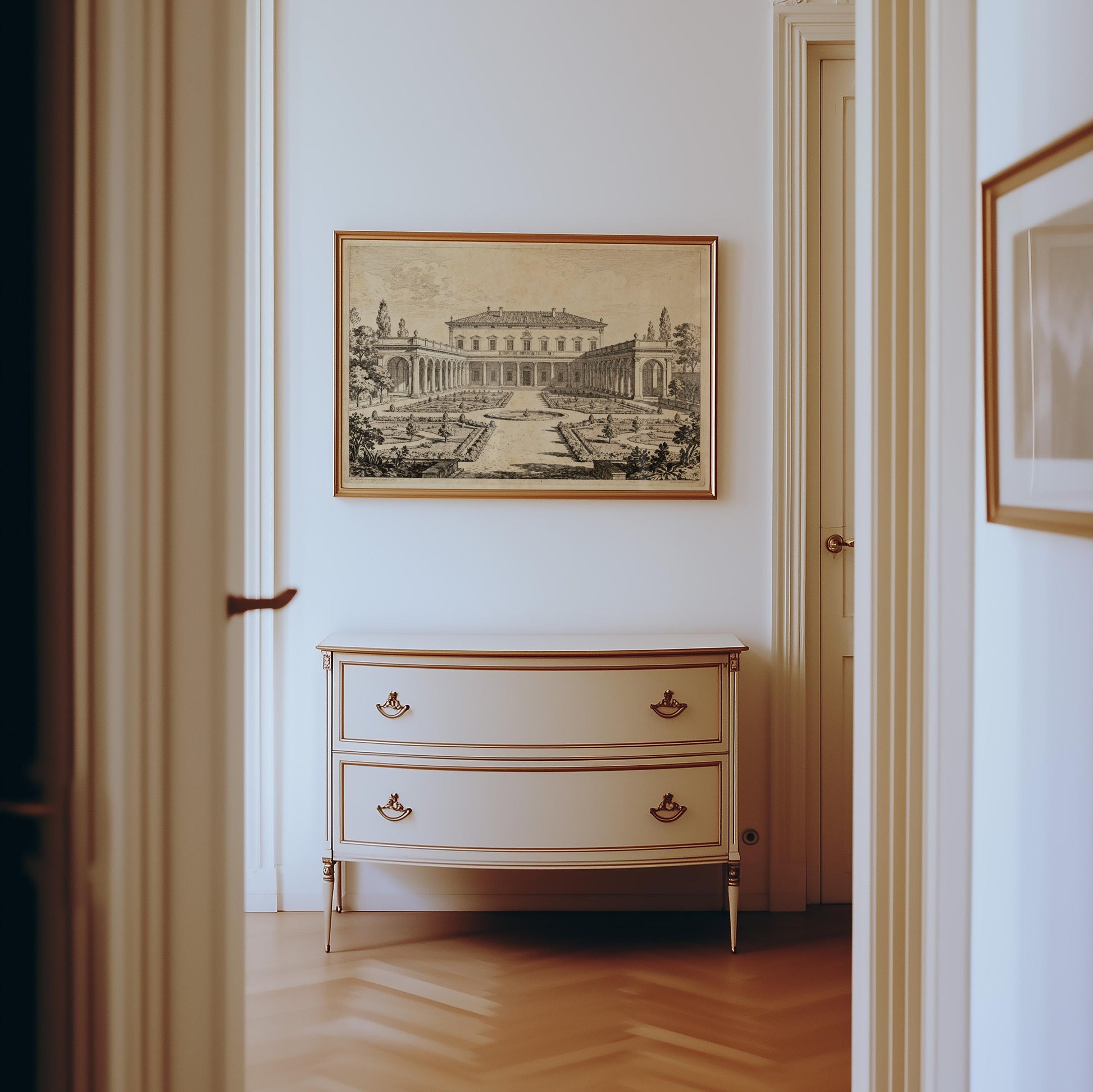 A hallway with wooden floors features a cream dresser with gold handles and an Italian Villa Art Print—Vintage Architectural Garden Courtyard from West & Wild, framed and hanging above it.