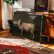 A Sheep Painting Print – Framed Vintage Country Home Artwork in Baroque Style leans against a wooden cabinet topped with a plant and artwork. Sunlight creates shadows on the red patterned rug and hardwood floor. Part of Vintage Art Prints & Framed Art.