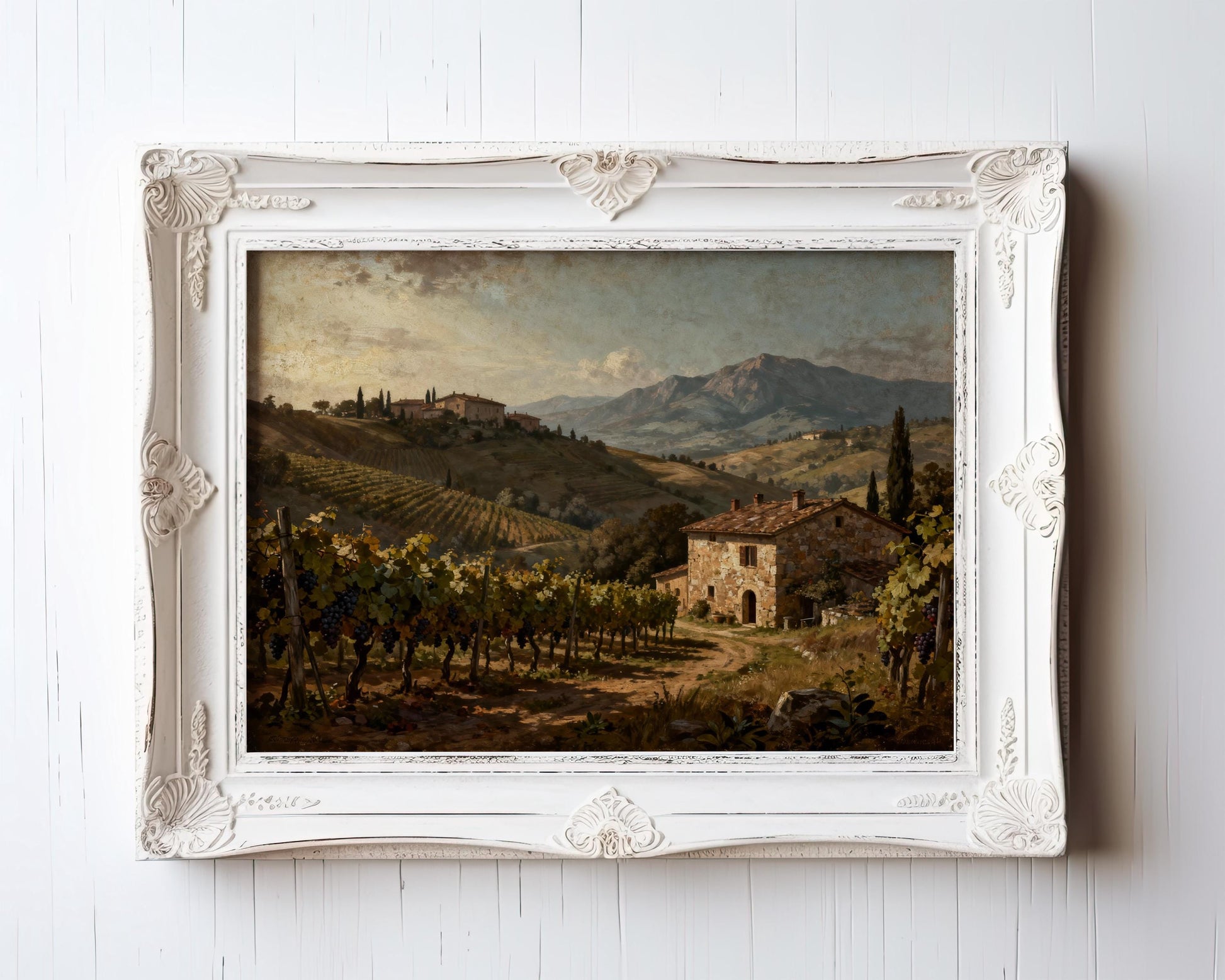 The Tuscan Vineyard Art Print – Framed Farmhouse Decor features a rustic stone house, cypress trees, and mountains under a cloudy sky. Its ornate white frame adds charm, making it ideal Mediterranean home decor.