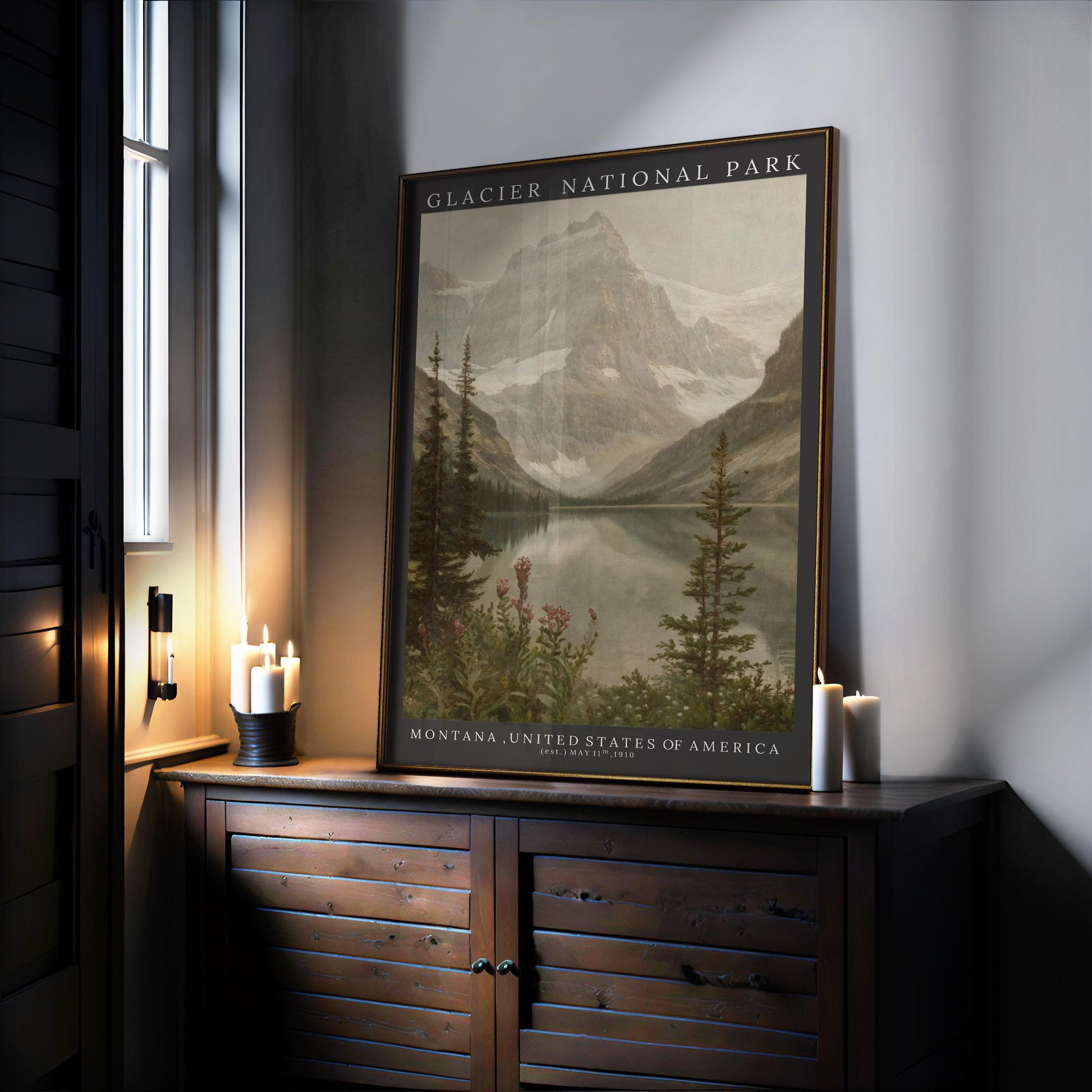A Glacier National Park Print – Vintage Mountain Valley Scene | Framed Nature Art sits on a dark wooden cabinet near candles and a window, displaying mountains, lake, and trees with “Montana, United States of America” below.