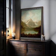 A Glacier National Park Poster – Framed Rustic Wall Art sits on a dark wooden cabinet by a window, surrounded by candles. This mountain lodge decor features mountains, trees, and a lake beneath a blue sky.