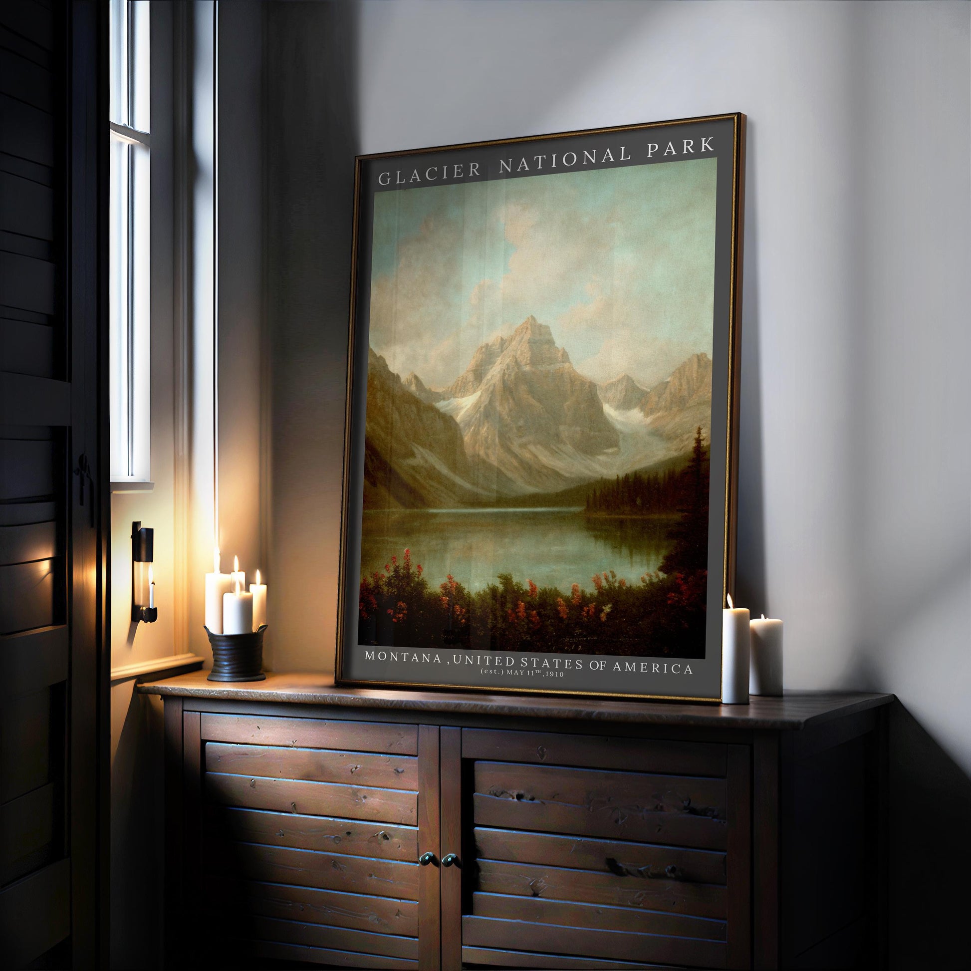 A Glacier National Park Poster – Framed Rustic Wall Art sits on a dark wooden cabinet by a window, surrounded by candles. This mountain lodge decor features mountains, trees, and a lake beneath a blue sky.