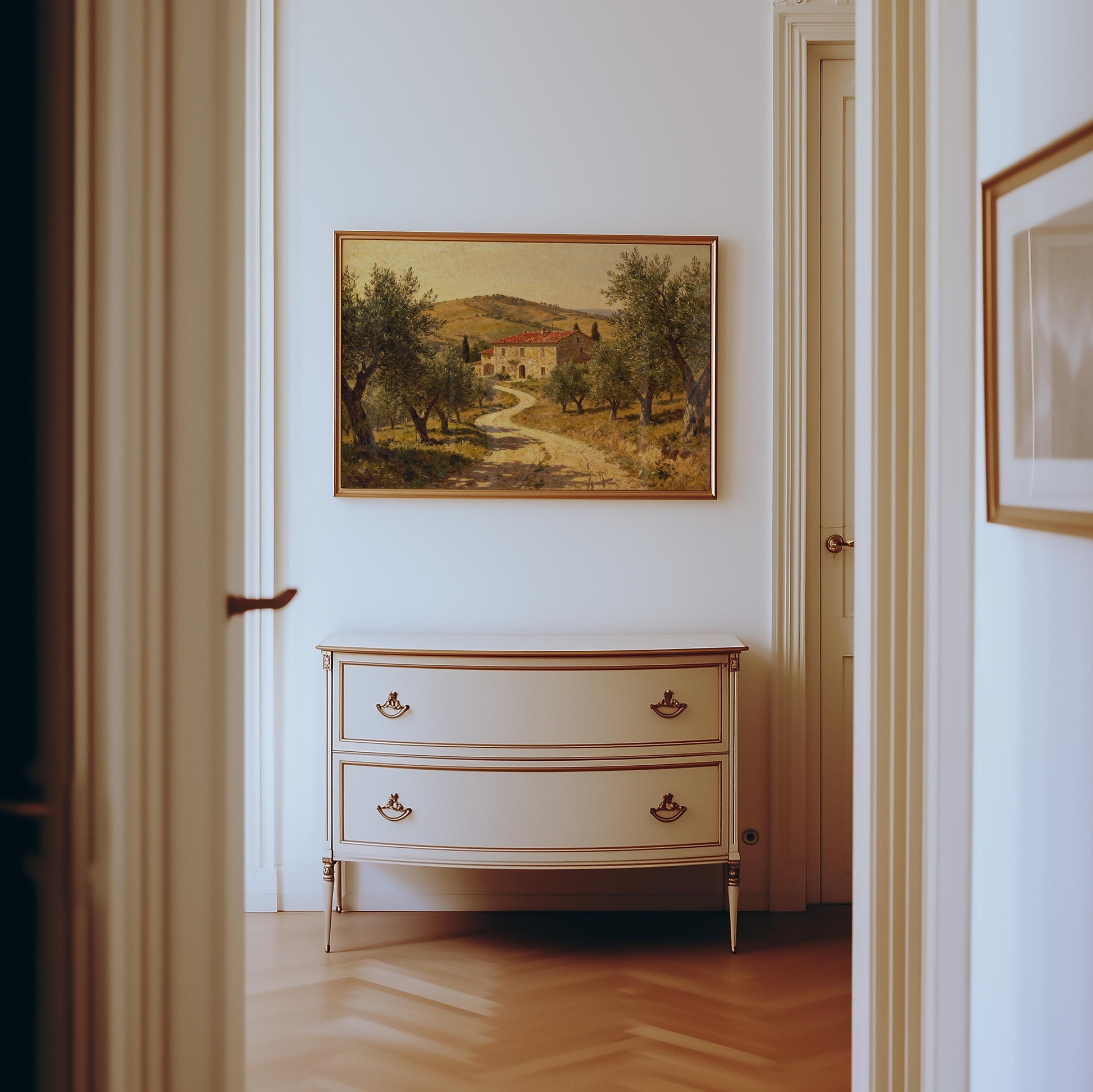 A hallway with light walls and wood floors features a cream dresser with two drawers, topped by a framed Tuscan Farmhouse Print—Vintage Olive Tree Art from West & Wild Art Archive—above it.
