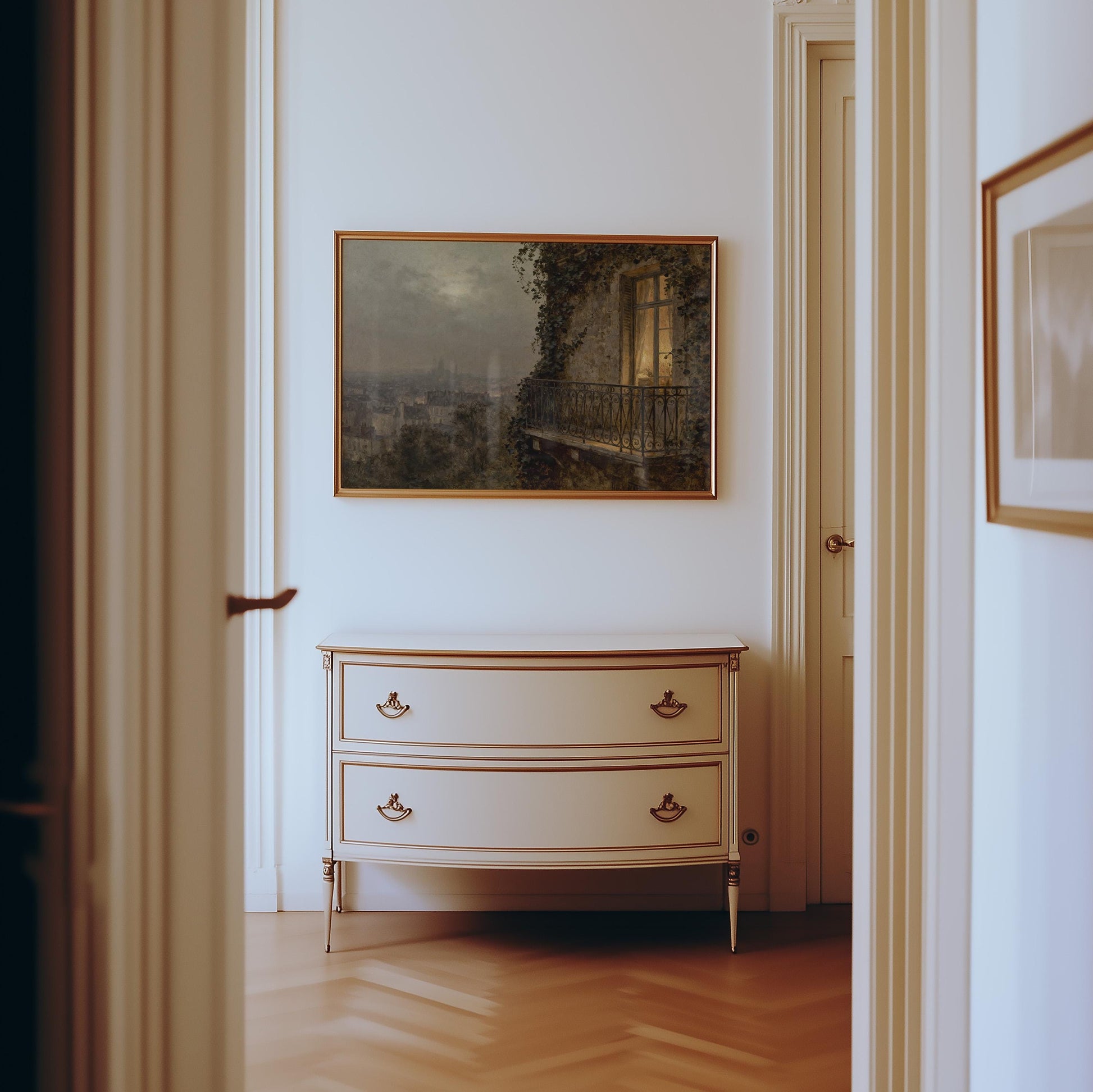 A white dresser with two drawers stands against a light wall. Above it hangs the Paris Balcony Art Print – Vintage Cityscape Artwork with Archival Matte Finish. The room features wooden floors and white door frames.