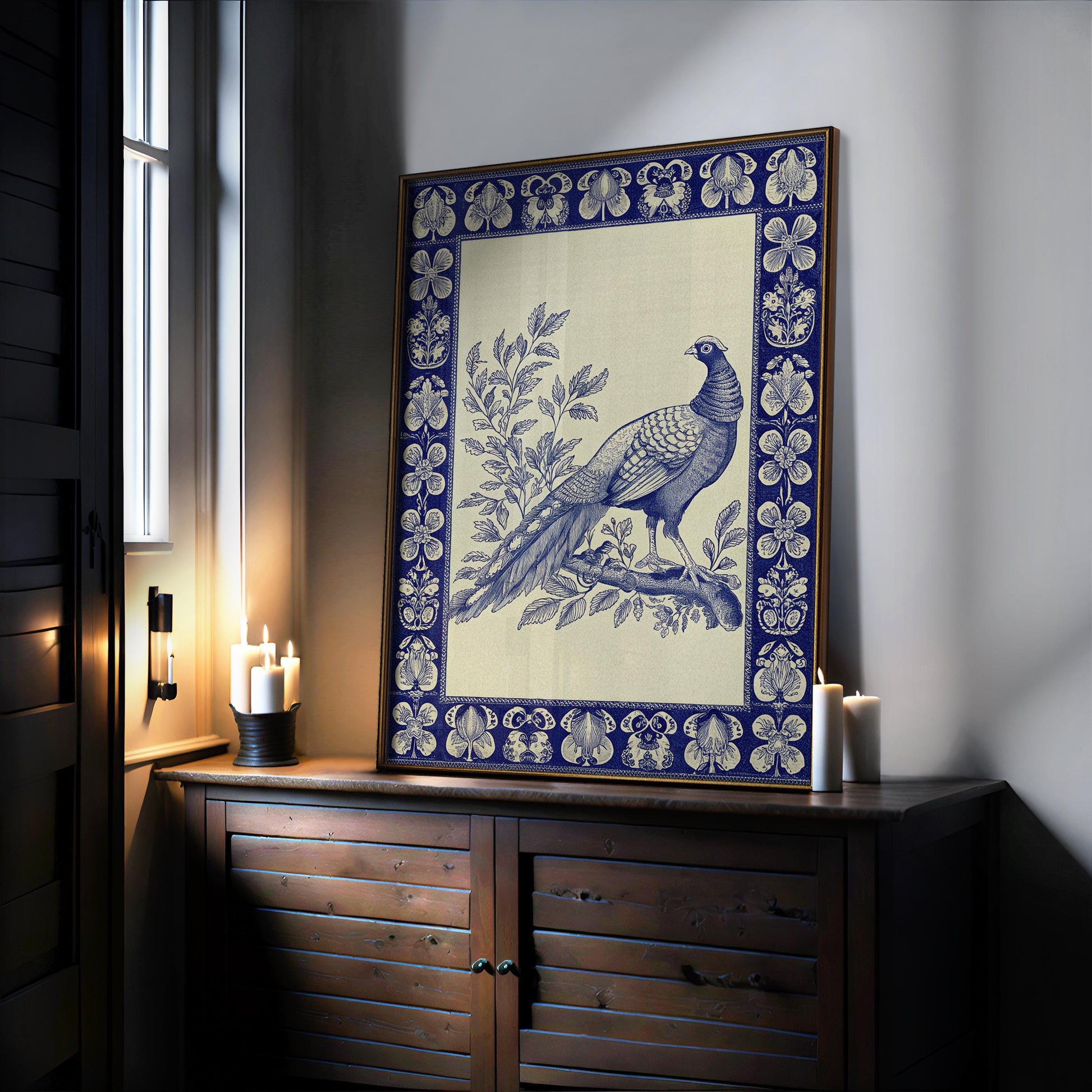 A Vintage Pheasant Wall Art print—an indigo bird on a branch with botanical details and a blue border in a wood-metal frame—rests on a cabinet by candles, softly lit, from the West & Wild Art Archive.