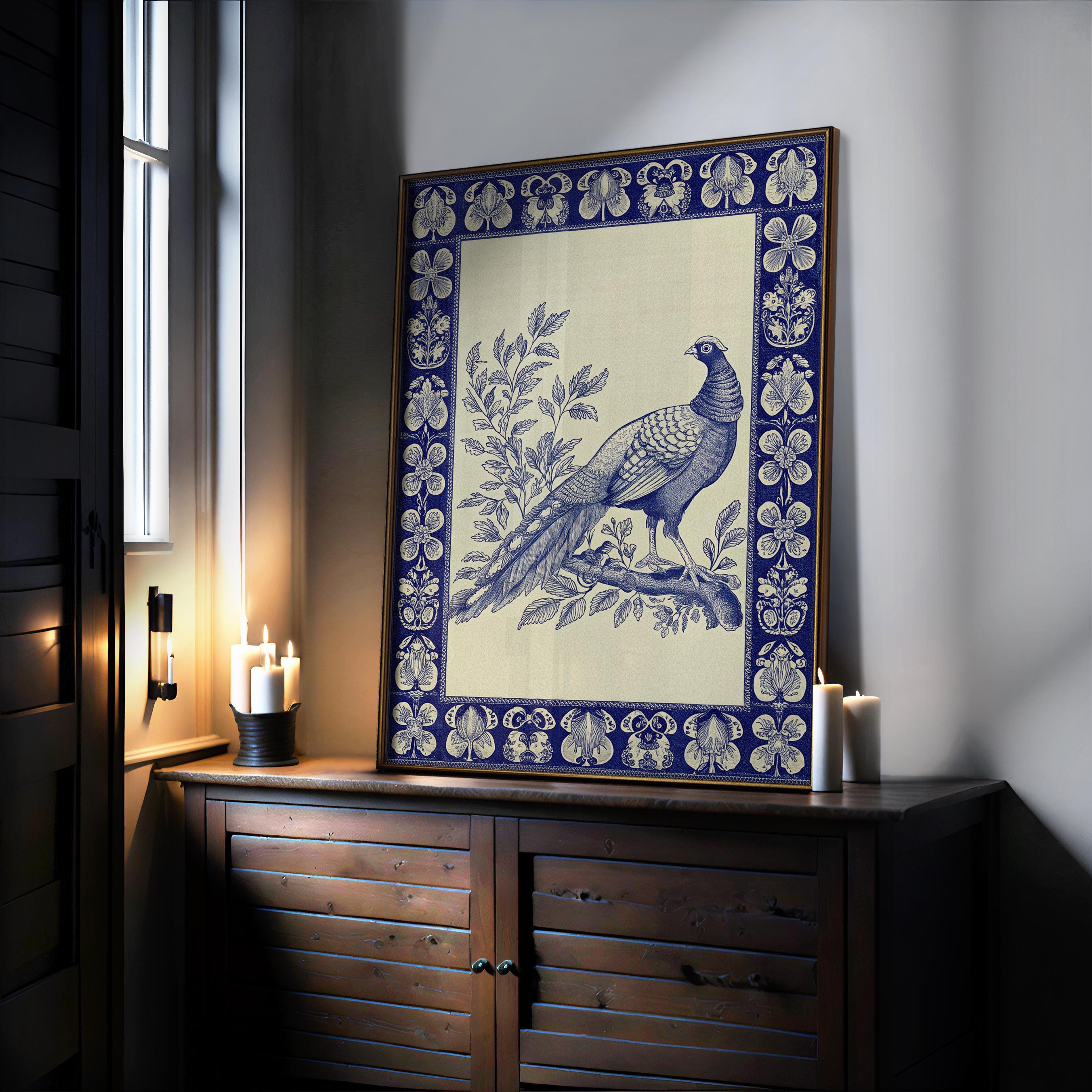 A Vintage Pheasant Wall Art print—an indigo bird on a branch with botanical details and a blue border in a wood-metal frame—rests on a cabinet by candles, softly lit, from the West & Wild Art Archive.