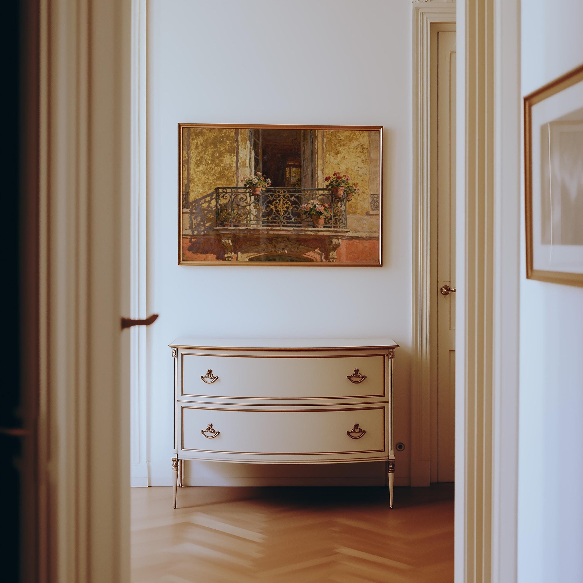 A white, vintage-style dresser with two drawers stands against a beige wall. Above it hangs the Vintage Paris Balcony Print – Golden Facade Art with Flower Pots from West & Wild, featuring doorways and a herringbone floor.
