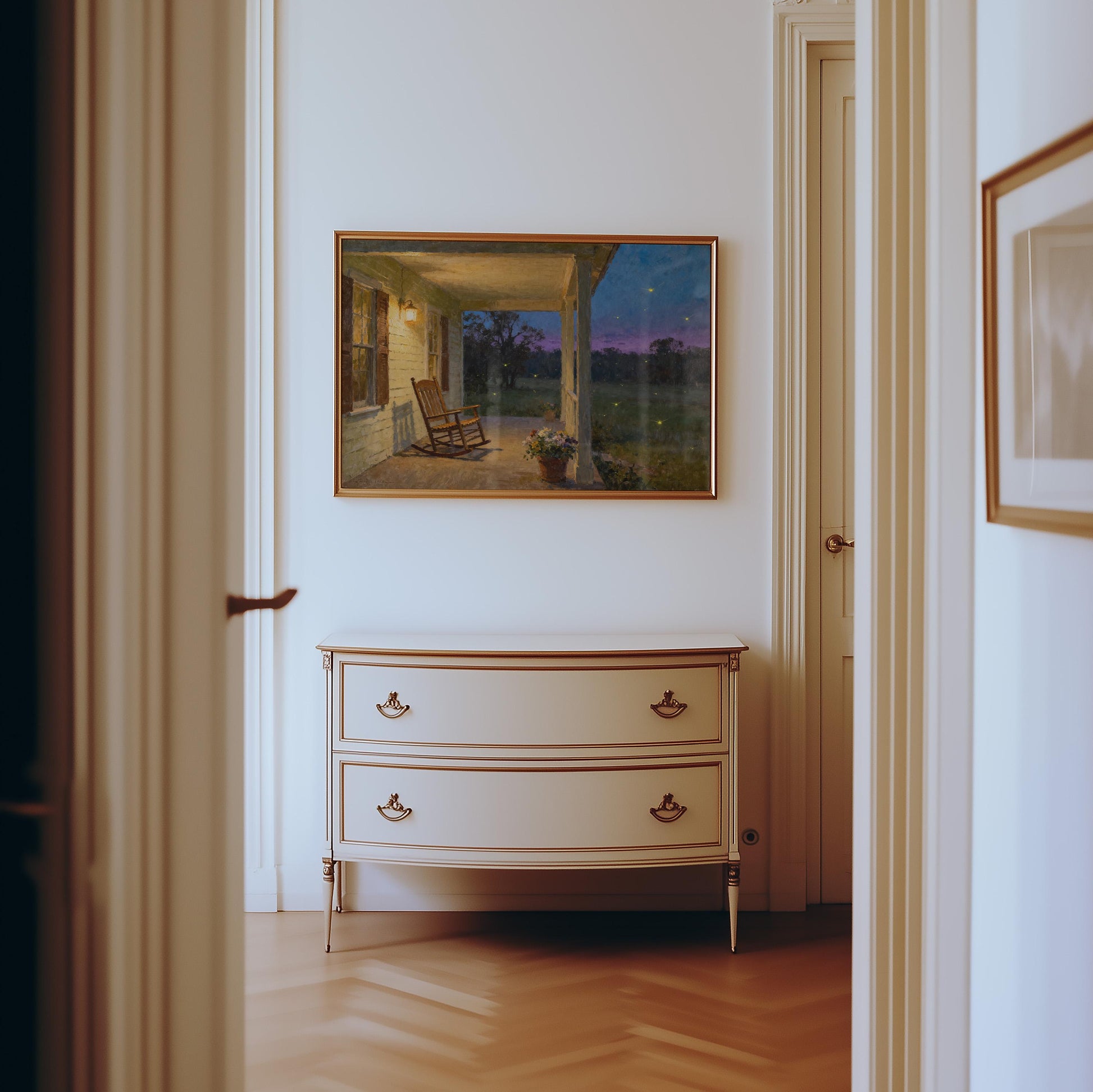 A hallway with light-colored walls and wooden floors features a cream-colored, two-drawer dresser beneath the Vintage Southern Porch Print – Rocking Chair Art with Fireflies from West & Wild Art Archive.