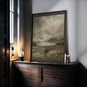 The Rocky Mountain Winter Print – Vintage Alpine Landscape, Framed Mountain Wall Art sits on a wooden cabinet by lit candles and a window, displaying snowy mountains and a lake beneath a cloudy sky.